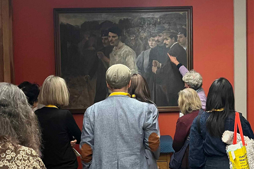 Photograph of man pointing to a figure in a painting with six people looking on.