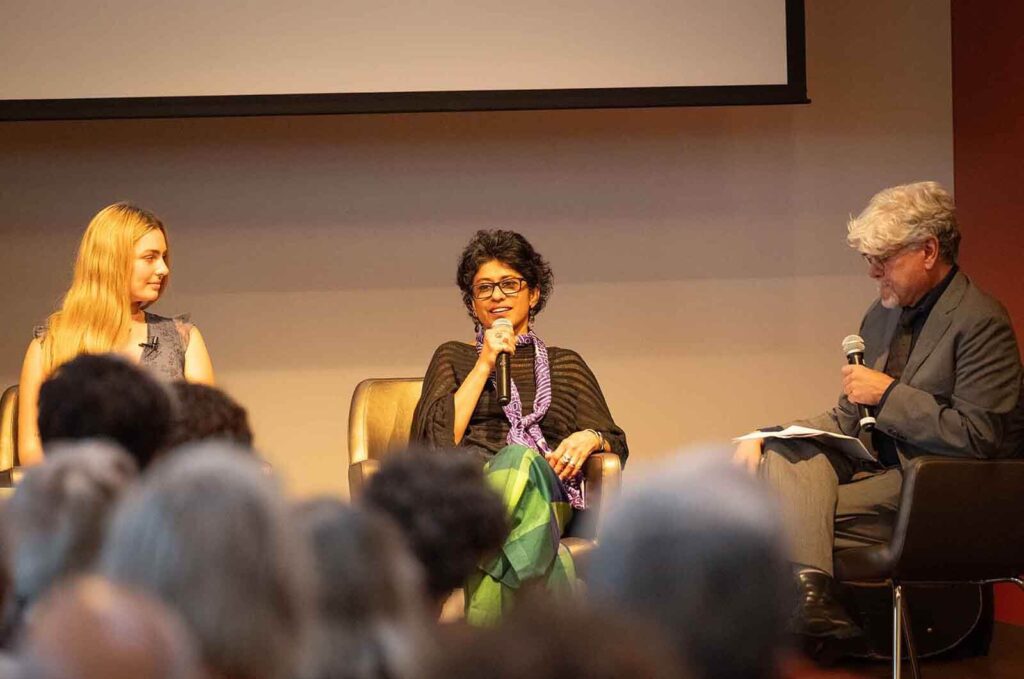 3 people on stage with woman in middle speaking to the audience